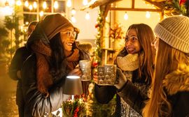 [Translate to Englisch:] Drei Frauen stehen an einem Glühweinstand auf einem Weihnachtsmarkt in München und trinken heißen Glühwein