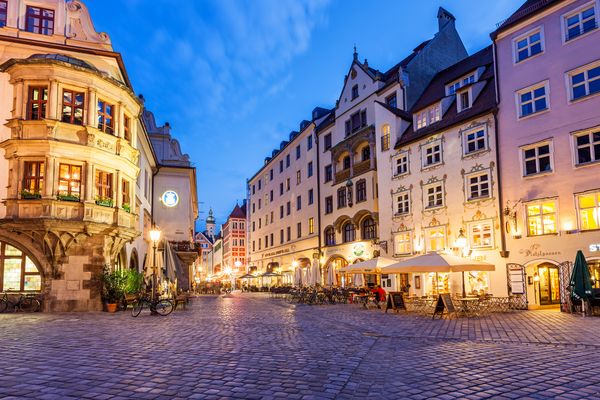 Evening view of Munich's Platzl with illuminated facades, cobblestone streets, and sidewalk cafés.