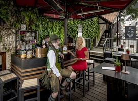 Ein Mann in bayrischer Tracht sitzt mit einer Dame in rotem Kleid an der Bar im Platzl Karree.