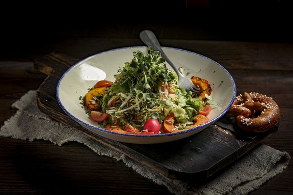 Ein liebevoll angerichteter Salat mit Gabel in einem weißen Teller begleitet von einer Brezel auf einem Servierbrett