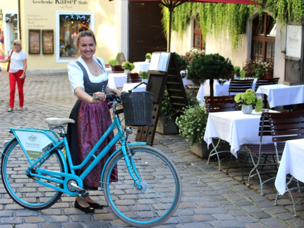 Frau in Dirndl posiert mit blauem Fahrrad vor einem Café mit gedeckten Tischen
