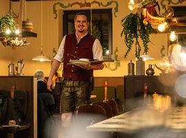 A waiter in a traditional outfit carries dishes, smiling inside a warmly lit restaurant with hanging plants.