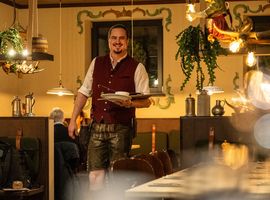 A waiter in a traditional outfit carries dishes, smiling inside a warmly lit restaurant with hanging plants.