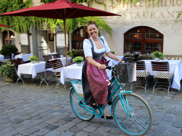 Frau in traditioneller Tracht fährt lächelnd auf einem blauen Fahrrad in einer gepflasterten Restaurantgasse