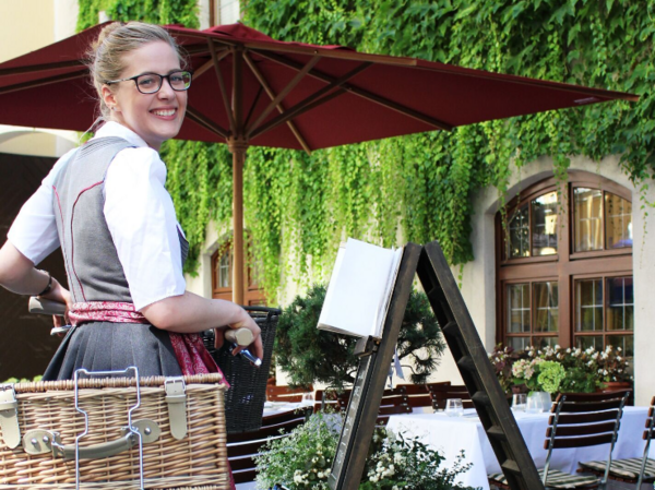 Lächelnde Frau in Dirndl-Tracht steht neben einem Fahrrad mit Weidenkorb vor einem begrünten Biergarten mit Tischen, Stühlen und einer roten Markise