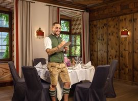 Ein Mitarbeiter der Platzl Hotels steht in bayerischer Tracht vor einem festlich in weiß gedecktem Tisch dunkelgrauen Stühlen.