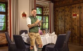 Ein Mitarbeiter der Platzl Hotels steht in bayerischer Tracht vor einem festlich in weiß gedecktem Tisch dunkelgrauen Stühlen.