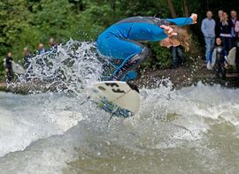 Ein Surfer in blauem Neoprenanzug an der Eisbachwelle vor Zuschauern.