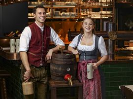 Zwei Mitarbeiter des Ayinger in der Au stehen in Tracht an der Bar an einem Bierfass angelehnt